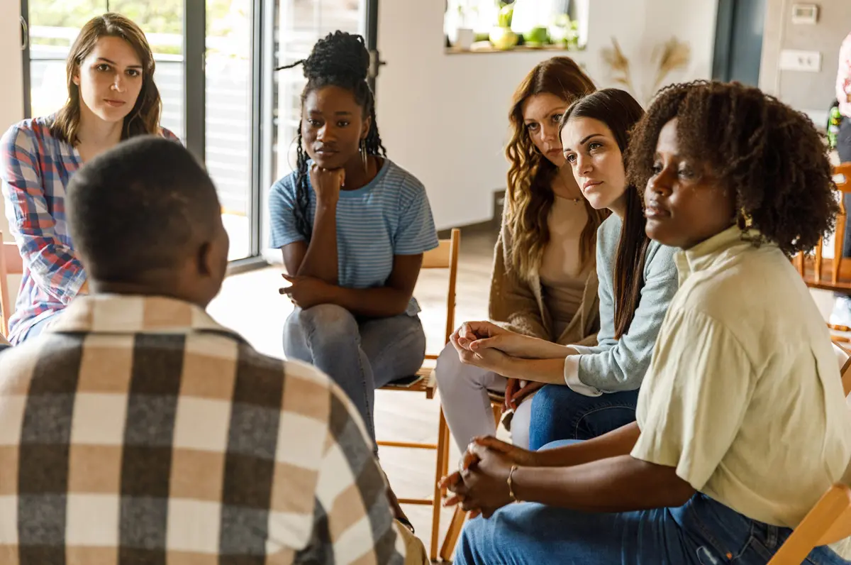A group of women participating in group therapy led by a partially unseen male therapist.
