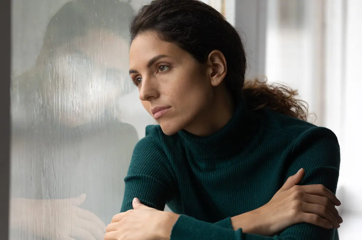 A depressed woman hugs herself while leaning her head against a window on a rainy day.